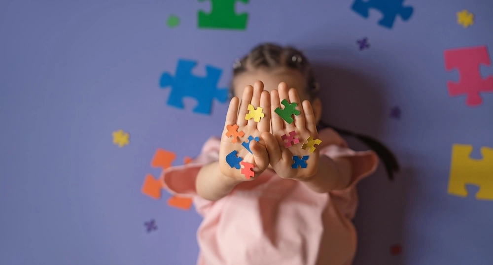 Bambina che mostra le mani dipinte davanti al viso, su fondo con tessere di puzzle